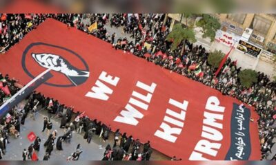 Aerial view of a protest with a large red banner bearing the words 'WE WILL KILL TRUMP' held by a crowd along a street. The banner features a black fist logo on the left.