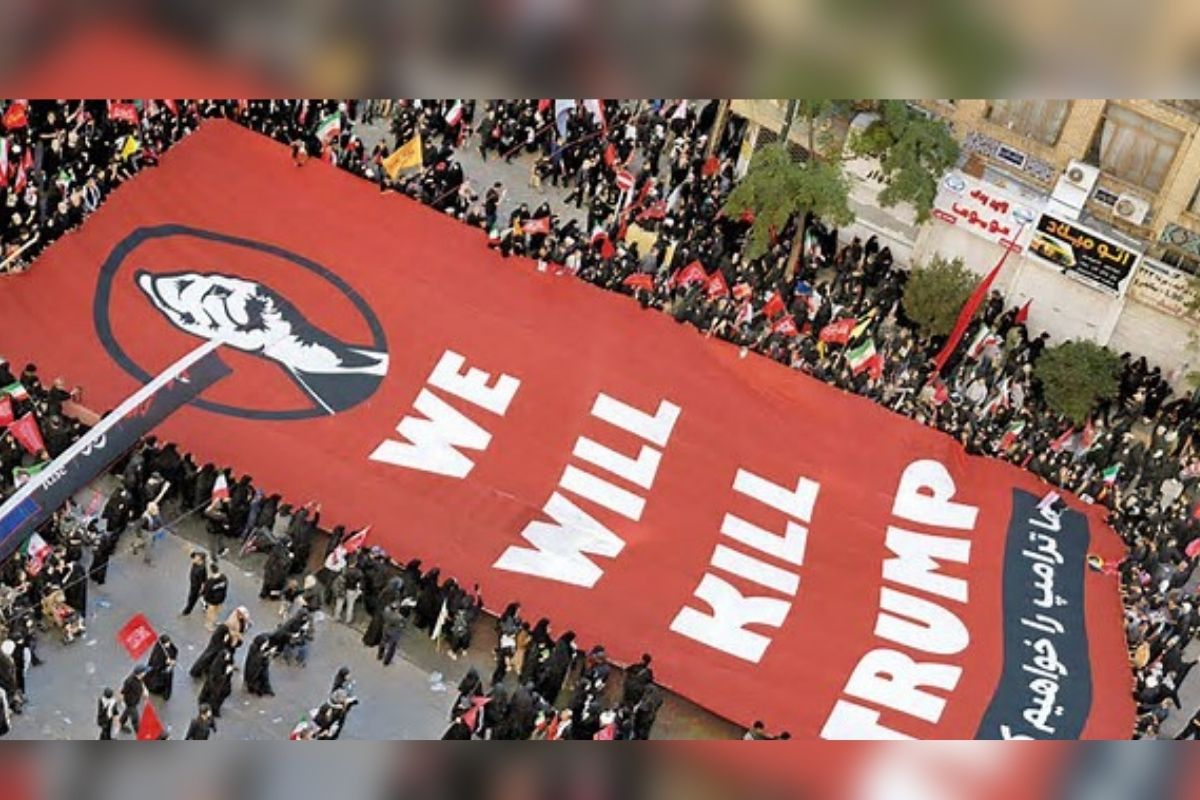 Aerial view of a protest with a large red banner bearing the words 'WE WILL KILL TRUMP' held by a crowd along a street. The banner features a black fist logo on the left.