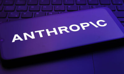 Smartphone screen showing the word ANTHROPIC in bold white letters, illuminated by a purple glow over a keyboard backdrop.