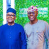 Two Nigerian men in traditional attire pose for a photo in front of a leafy green backdrop; one wears a dark blue agbada and cap, the other a light gray outfit with a patterned cap, both smiling.