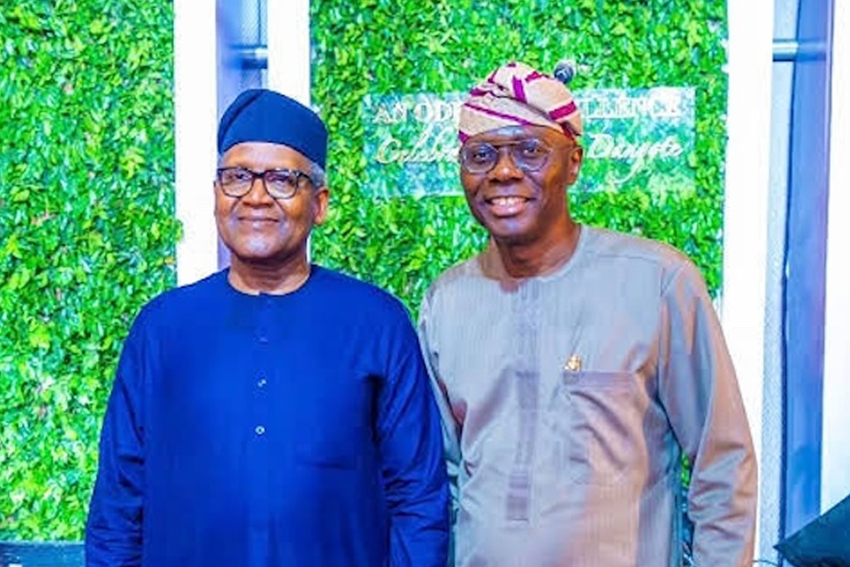Two Nigerian men in traditional attire pose for a photo in front of a leafy green backdrop; one wears a dark blue agbada and cap, the other a light gray outfit with a patterned cap, both smiling.