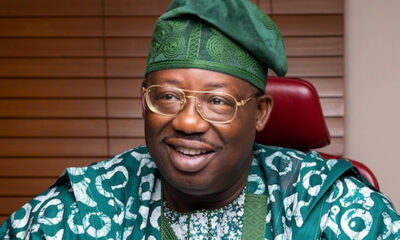 Smiling man in green traditional outfit and cap, seated indoors with wooden blinds behind.