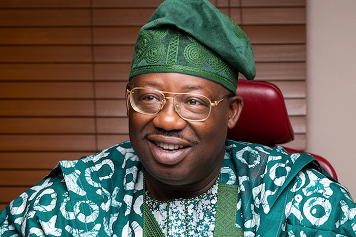 Smiling man in green traditional outfit and cap, seated indoors with wooden blinds behind.