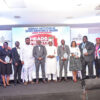 Group of professionals in suits and dresses posing with glass awards on a stage during an awards ceremony.