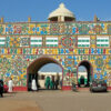 Colorful decorative archway with two arches covered in bright floral patterns; people walking and cars parked in front.