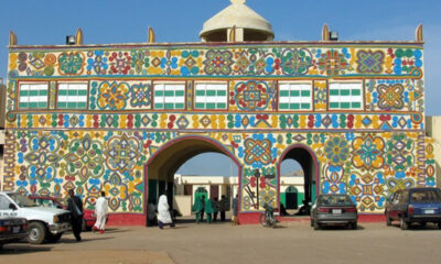 Colorful decorative archway with two arches covered in bright floral patterns; people walking and cars parked in front.