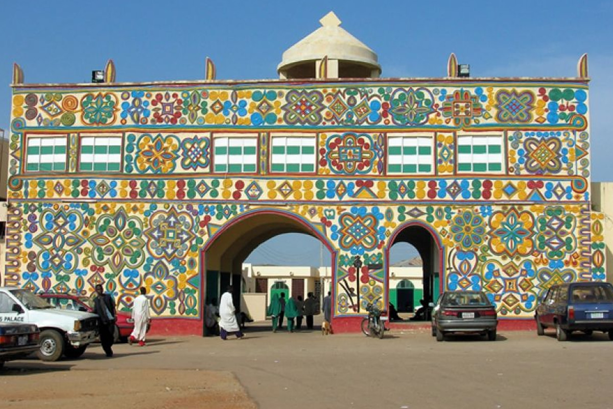 Colorful decorative archway with two arches covered in bright floral patterns; people walking and cars parked in front.