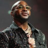 Hip-hop artist on stage wearing a studded black shirt, gold chains, and glasses, looking upward while performing.
