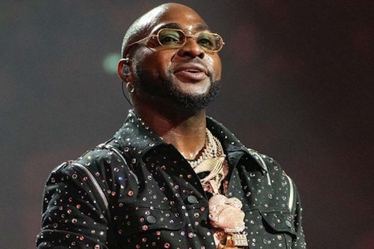 Hip-hop artist on stage wearing a studded black shirt, gold chains, and glasses, looking upward while performing.