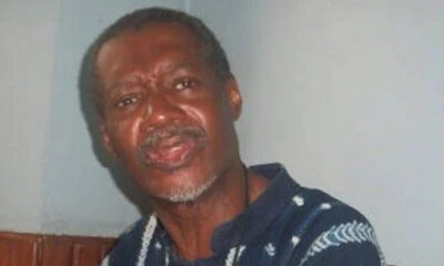 Older man with gray beard in a dark blue patterned shirt, looking toward the camera in an indoor setting.