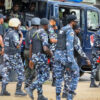 Group of armed police and soldiers near a blue armored vehicle, some in camouflage uniforms and helmets, preparing to mobilize.