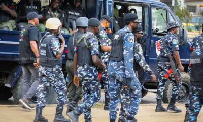 Group of armed police and soldiers near a blue armored vehicle, some in camouflage uniforms and helmets, preparing to mobilize.