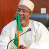 Older man in white traditional attire and cap sits at a desk with a green flag and a microphone in front of him, in a formal setting.