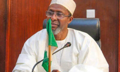 Older man in white traditional attire and cap sits at a desk with a green flag and a microphone in front of him, in a formal setting.