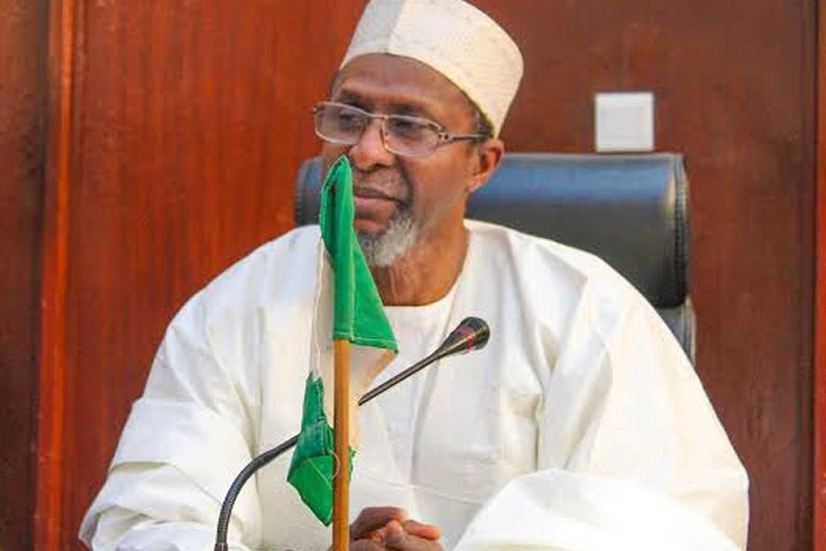 Older man in white traditional attire and cap sits at a desk with a green flag and a microphone in front of him, in a formal setting.