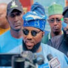 Man in blue traditional attire wearing blue headwrap and sunglasses speaks to reporters amid a press crowd, microphones in front of him