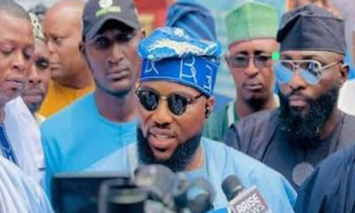 Man in blue traditional attire wearing blue headwrap and sunglasses speaks to reporters amid a press crowd, microphones in front of him