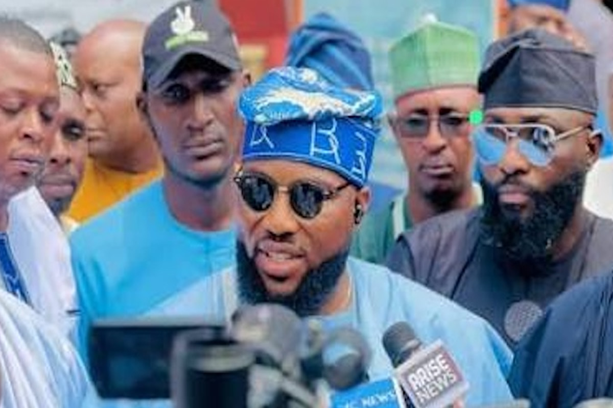 Man in blue traditional attire wearing blue headwrap and sunglasses speaks to reporters amid a press crowd, microphones in front of him