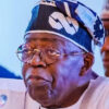 Elderly man with round glasses and a blue patterned traditional cap speaking at a meeting.