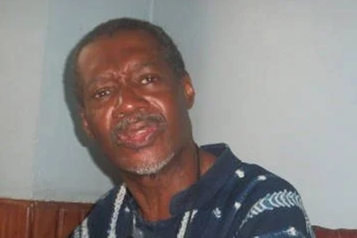 Older man with gray beard in a dark blue patterned shirt, looking toward the camera in an indoor setting.