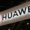Huawei logo and wordmark on a white illuminated sign hanging indoors, background of ceiling lights