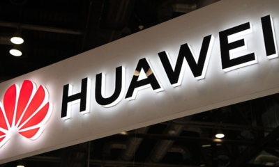 Huawei logo and wordmark on a white illuminated sign hanging indoors, background of ceiling lights