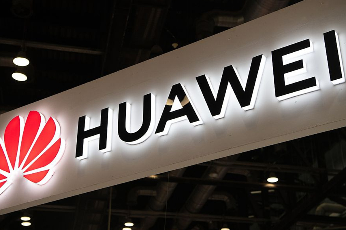 Huawei logo and wordmark on a white illuminated sign hanging indoors, background of ceiling lights