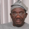 Man wearing a light gray traditional outfit and patterned headwrap, looking at the camera with a neutral expression while wearing glasses
