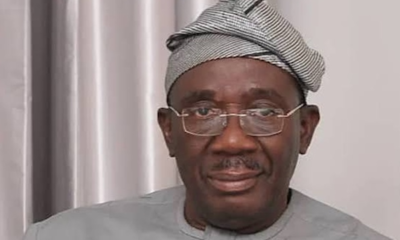 Man wearing a light gray traditional outfit and patterned headwrap, looking at the camera with a neutral expression while wearing glasses