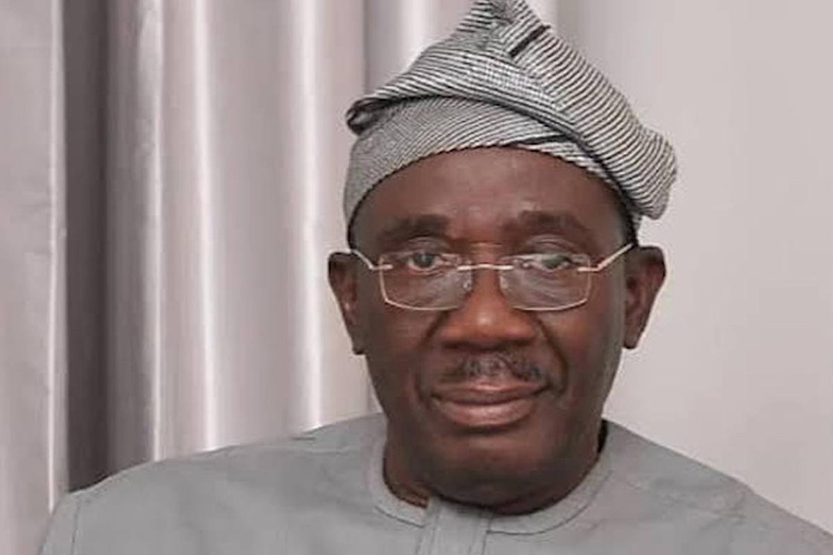 Man wearing a light gray traditional outfit and patterned headwrap, looking at the camera with a neutral expression while wearing glasses