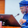 Man in a blue traditional agbada and cap speaks at a wooden lectern with a microphone and glasses, reading from notes.