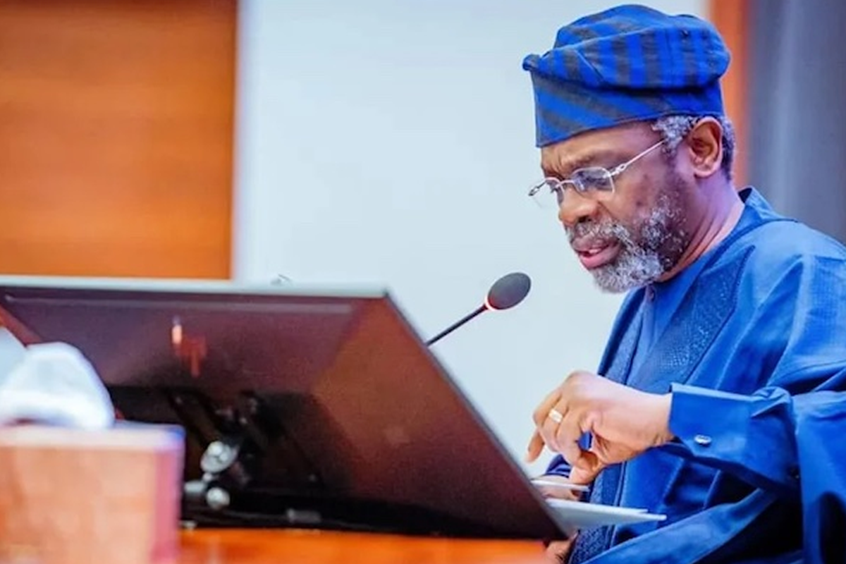 Man in a blue traditional agbada and cap speaks at a wooden lectern with a microphone and glasses, reading from notes.