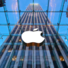 White Apple logo displayed on the glass facade of a tall modern building against a blue sky. The city skyline is reflected in the glass around it.