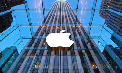White Apple logo displayed on the glass facade of a tall modern building against a blue sky. The city skyline is reflected in the glass around it.