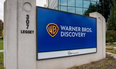 Outdoor sign at 307 Legget reading 'Warner Bros. Discovery' with WB shield logo on a blue panel at a corporate entrance site.