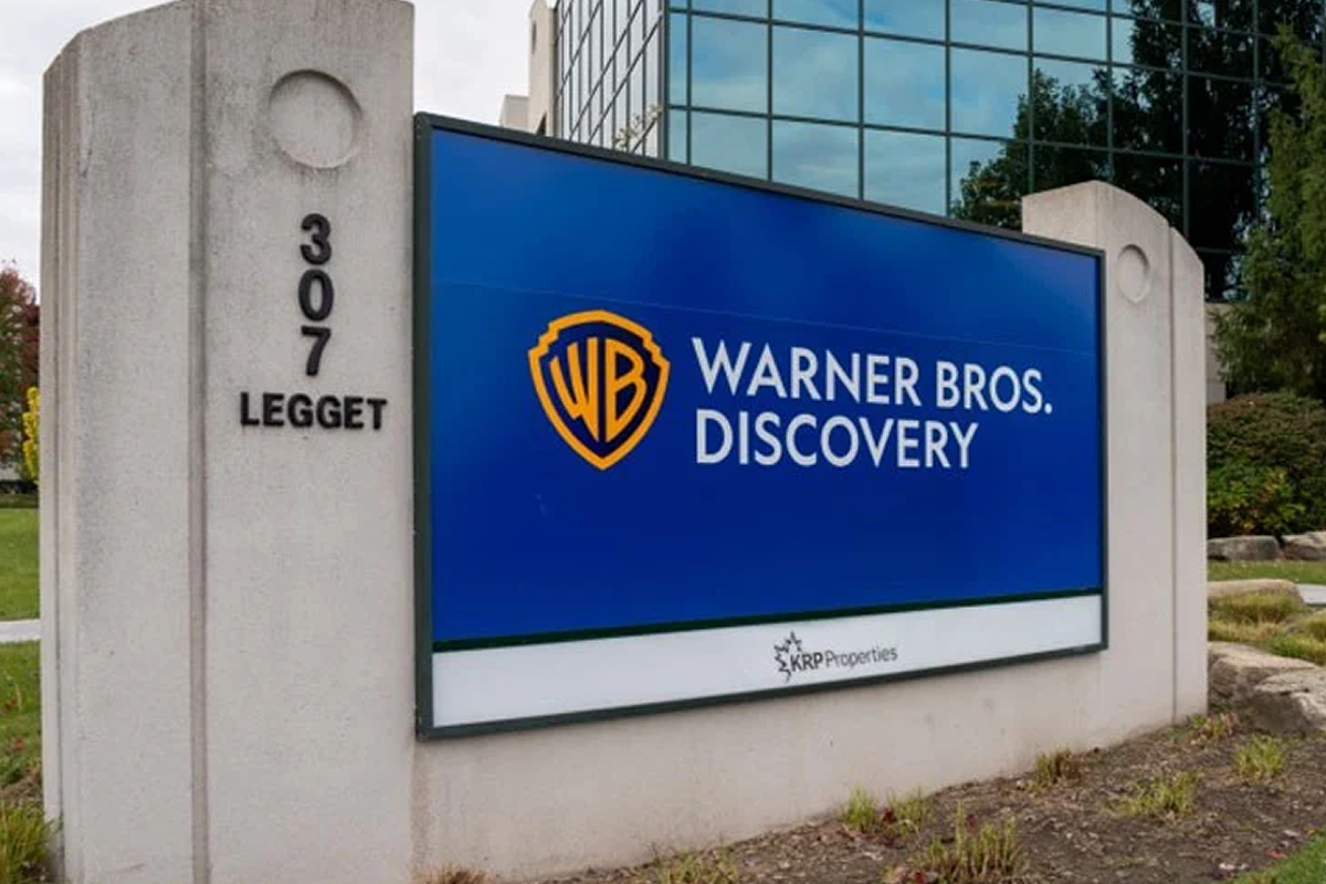 Outdoor sign at 307 Legget reading 'Warner Bros. Discovery' with WB shield logo on a blue panel at a corporate entrance site.