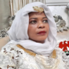 Woman in a white hijab and ornate gown with gold trim, seated on a silver throne-like chair with red eagle embroidery on the cushion.