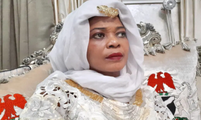 Woman in a white hijab and ornate gown with gold trim, seated on a silver throne-like chair with red eagle embroidery on the cushion.