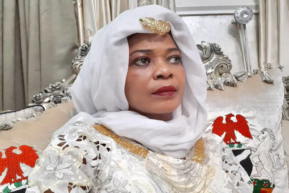 Woman in a white hijab and ornate gown with gold trim, seated on a silver throne-like chair with red eagle embroidery on the cushion.