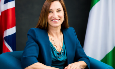 Professional portrait of a smiling woman in a teal blouse and blue blazer, with flags in the background.