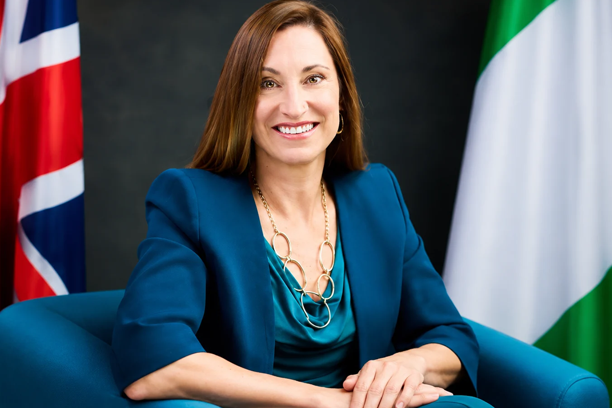 Professional portrait of a smiling woman in a teal blouse and blue blazer, with flags in the background.