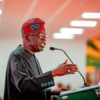 Speaker wearing a red headwrap and dark traditional outfit speaks into a microphone at a green podium.