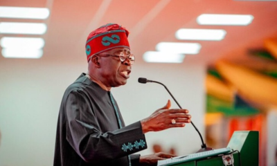 Speaker wearing a red headwrap and dark traditional outfit speaks into a microphone at a green podium.