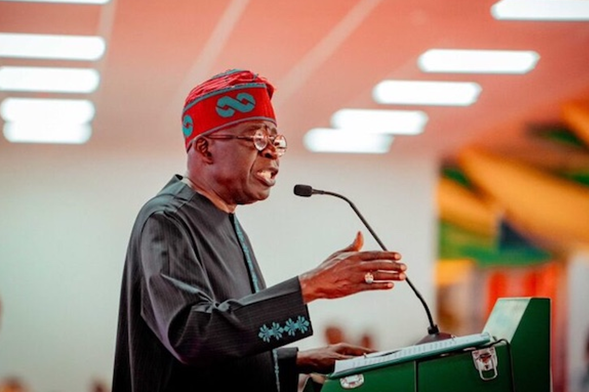 Speaker wearing a red headwrap and dark traditional outfit speaks into a microphone at a green podium.