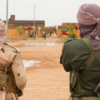 Two armed soldiers stand with their backs to the camera in a dusty town, wearing camouflage uniforms and wrapped faces, a rifle slung over one shoulder.