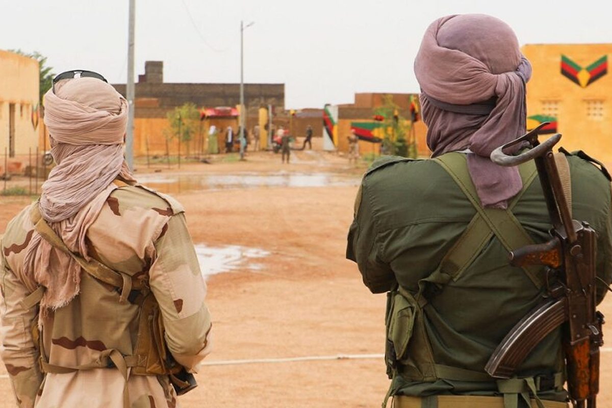 Two armed soldiers stand with their backs to the camera in a dusty town, wearing camouflage uniforms and wrapped faces, a rifle slung over one shoulder.
