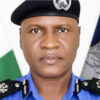 Portrait of a police officer in a blue uniform and black beret, with a flag in the background representing service and duty.