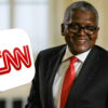 Smiling man in a dark suit and red tie with a large CNN logo foreground.