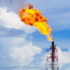 Industrial flare stack emitting a large orange flame into a blue sky with white clouds, at a processing facility