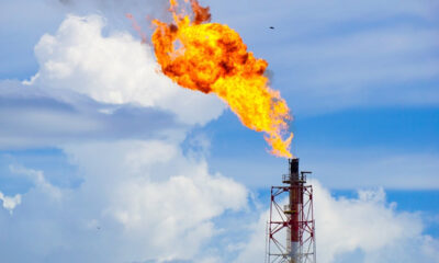 Industrial flare stack emitting a large orange flame into a blue sky with white clouds, at a processing facility